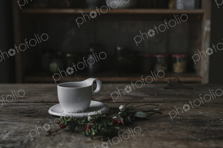 A white china cup and saucer on a kitchen table and a twig of pine and berries.