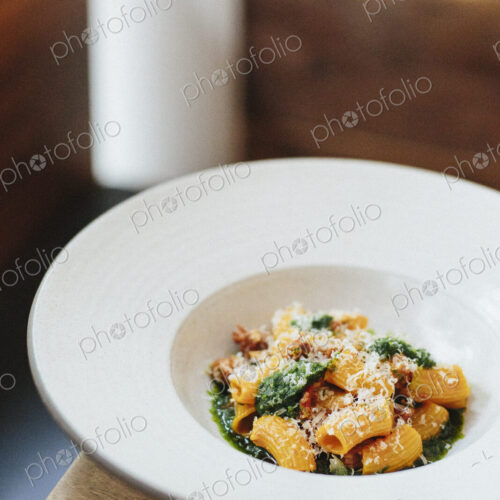 A white dish with pasta and a vegetable sauce and shavings of parmesan.