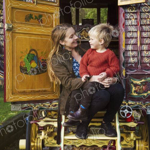 A woman and a small boy seated on the steps of a bow top caravan painted in a…