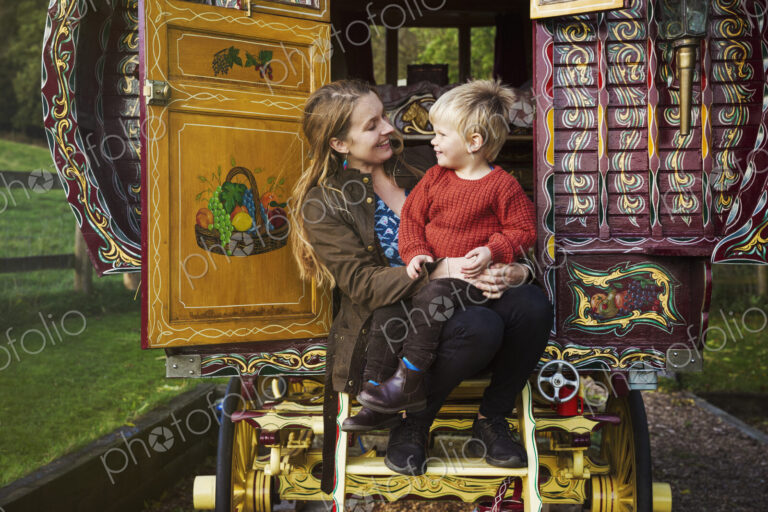A woman and a small boy seated on the steps of a bow top caravan painted in a…