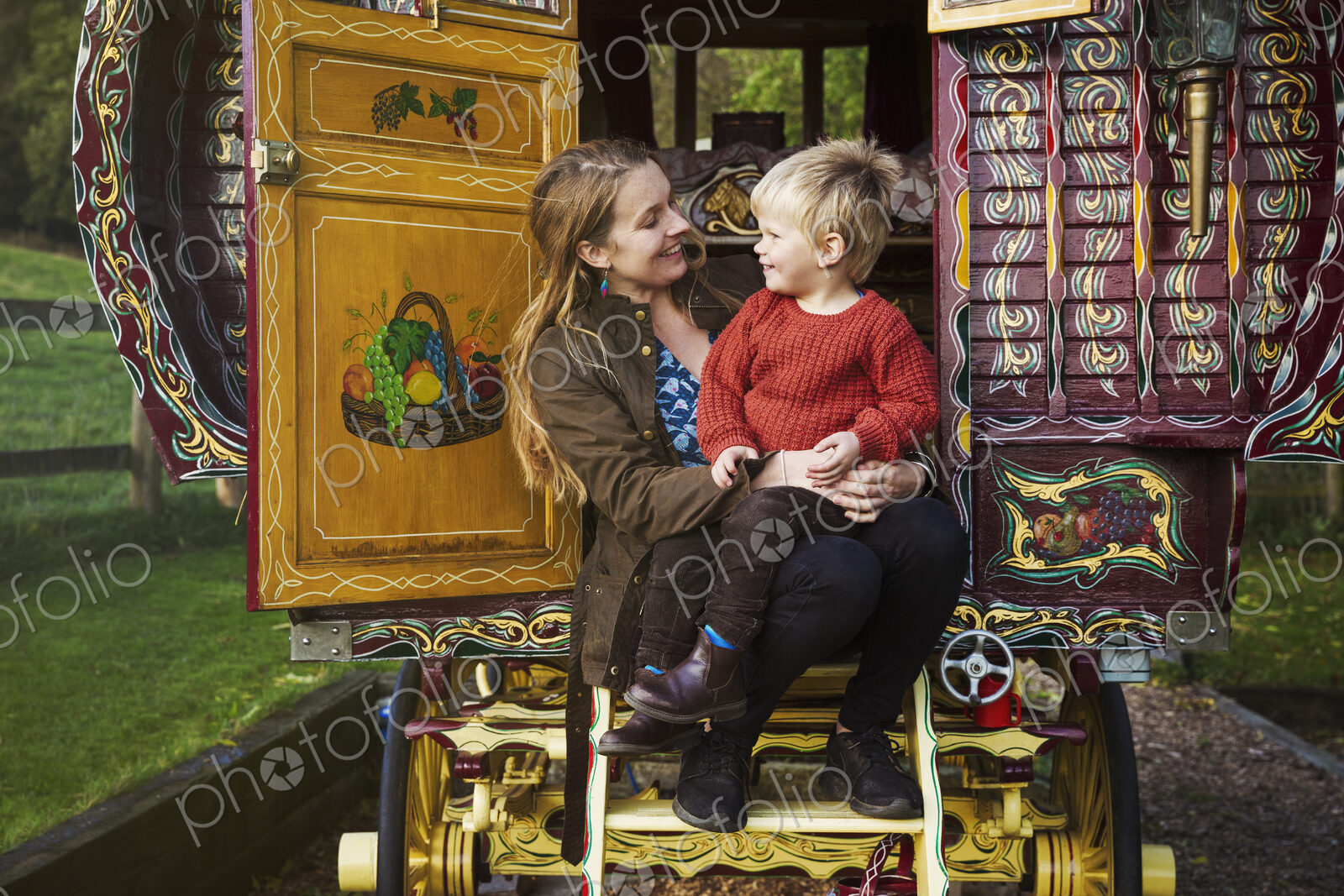 A woman and a small boy seated on the steps of a bow top caravan painted in a…