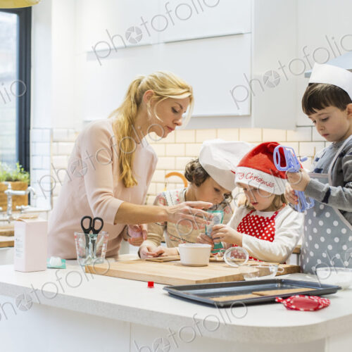 A woman and three children working together, making a gingerbread house, and icing the…