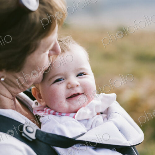 A woman carrying a baby in a baby carrier, kissing her on the head.