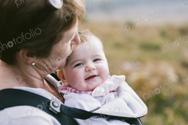 A woman carrying a baby in a baby carrier, kissing her on the head.
