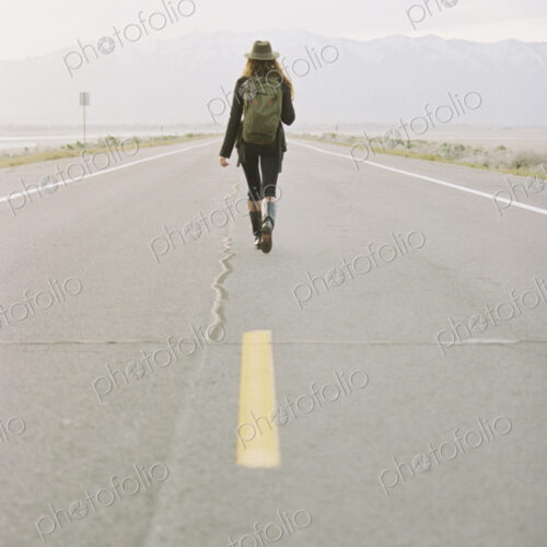 A woman carrying a backpack walking down the centre line of a country road.