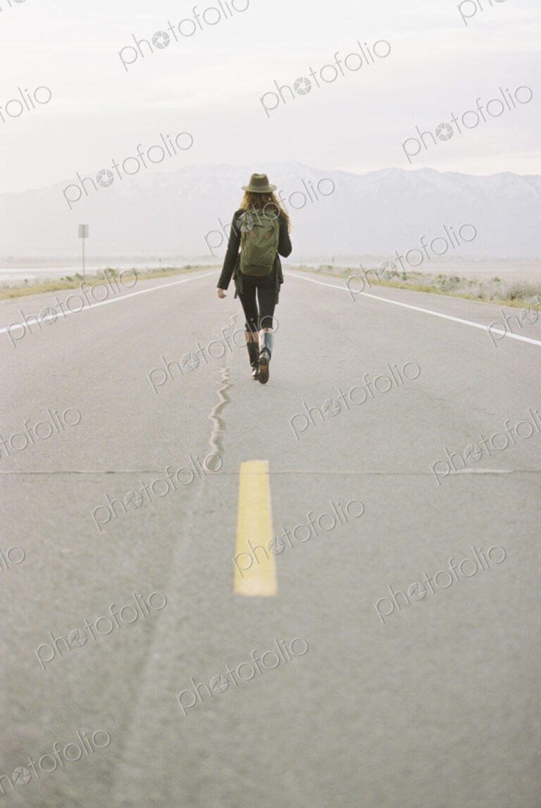 A woman carrying a backpack walking down the centre line of a country road.