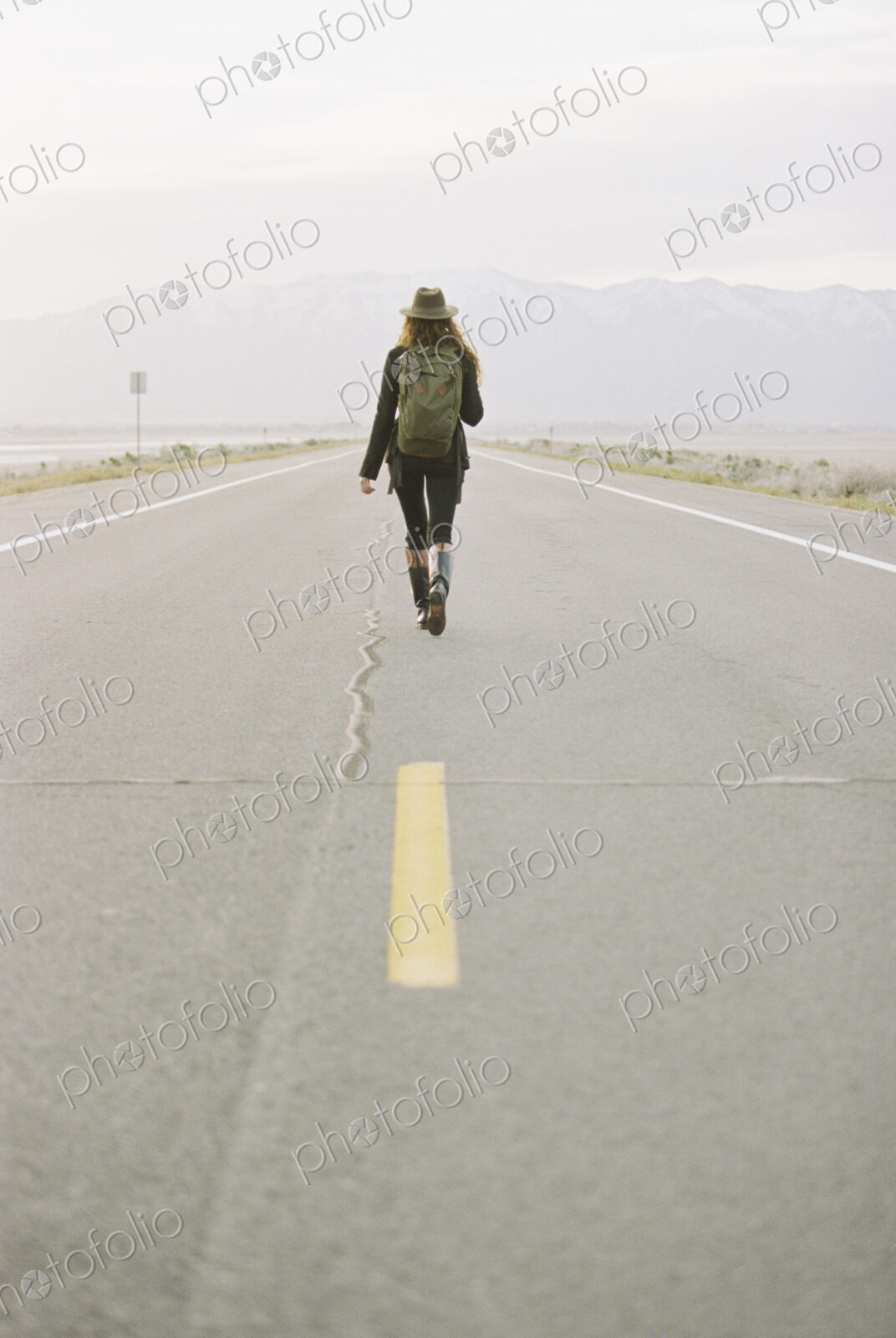 A woman carrying a backpack walking down the centre line of a country road.