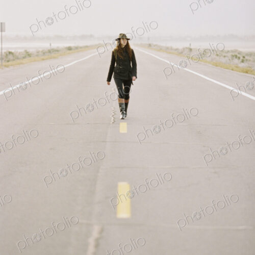 A woman carrying a backpack walking down the centre line of a country road.
