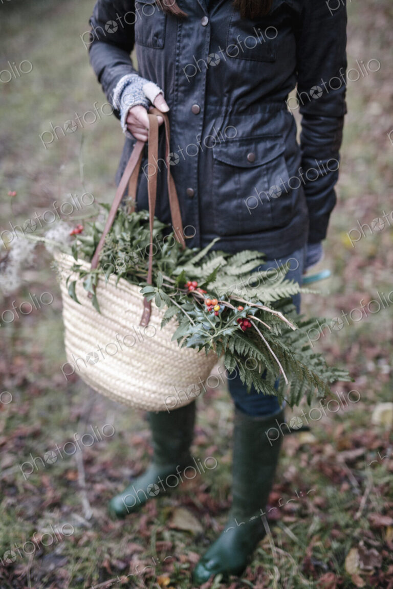 A woman carrying a basket of foliage, evergreen sprigs and glossy laurel with berries.