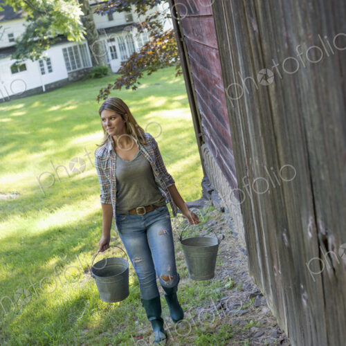 A woman carrying a bucket for animal feed in each hand, outside a barn.