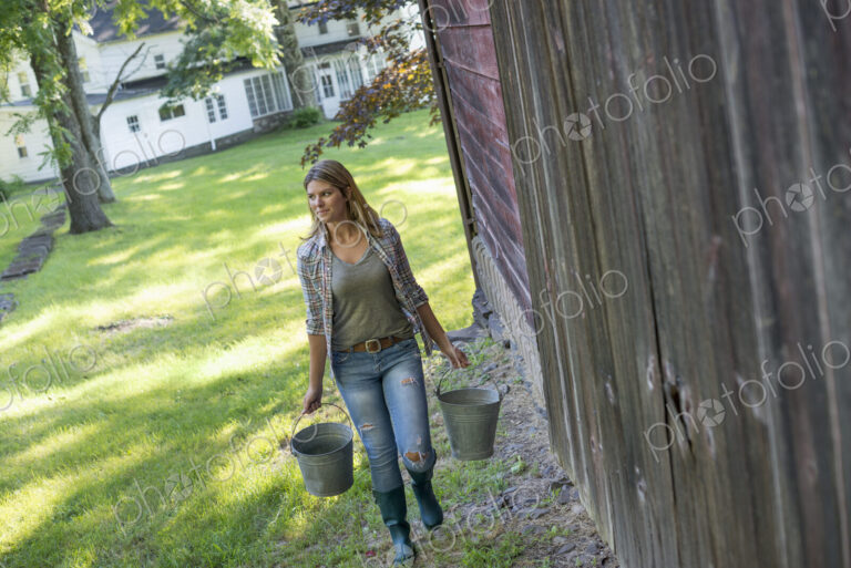A woman carrying a bucket for animal feed in each hand, outside a barn.