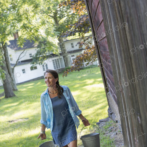 A woman carrying a bucket for animal feed in each hand, outside a barn.