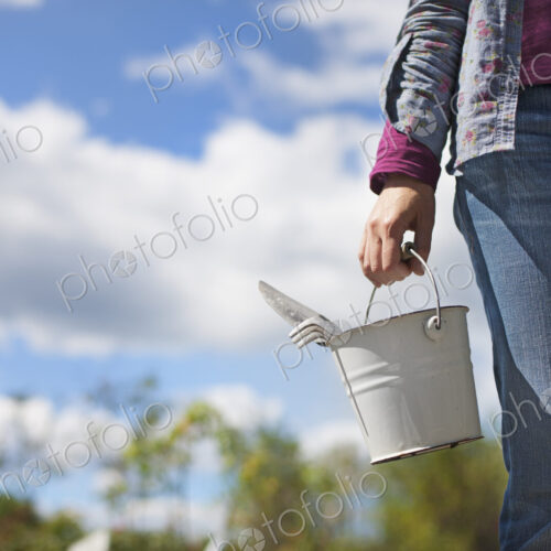 A woman carrying a white tin bucket with gardening tools, on an organic farm.