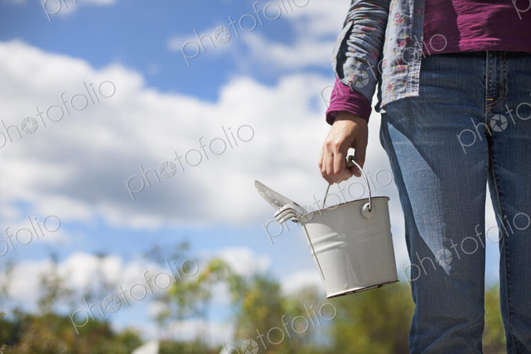 A woman carrying a white tin bucket with gardening tools, on an organic farm.