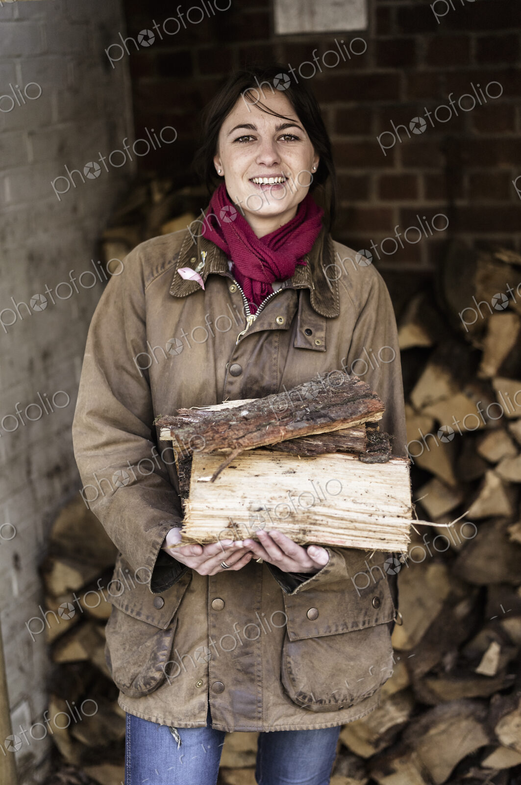 A woman carrying split logs for firewood from a large log store.
