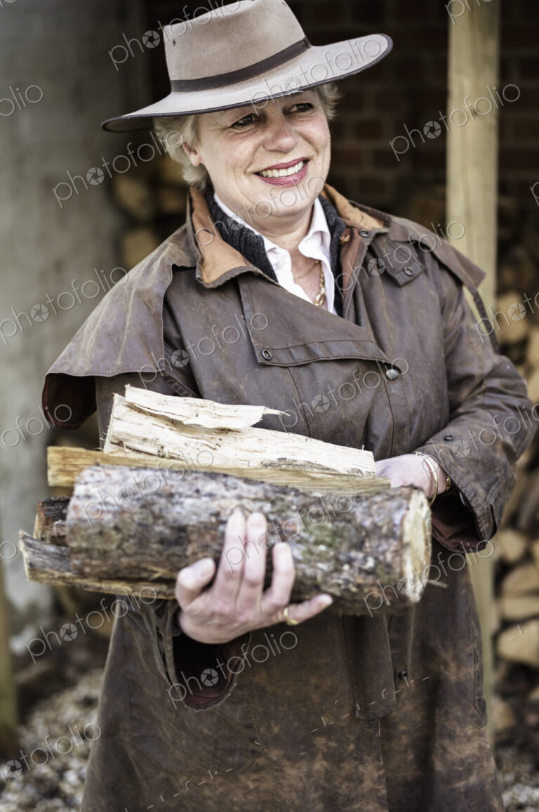 A woman carrying split logs for firewood from a large log store.