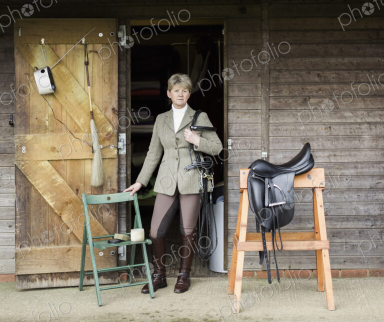 A woman cleaning and preparing tack and saddle, in the courtyard of a riding stable.