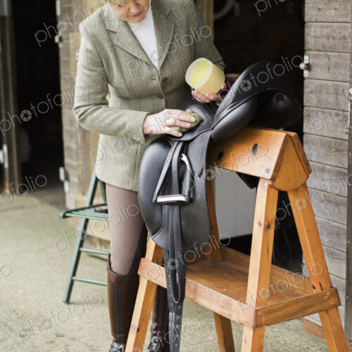 A woman cleaning and preparing tack and saddle, in the courtyard of a riding stable. A woman cleaning and preparing tack and saddle, in the courtyard of a riding stable.