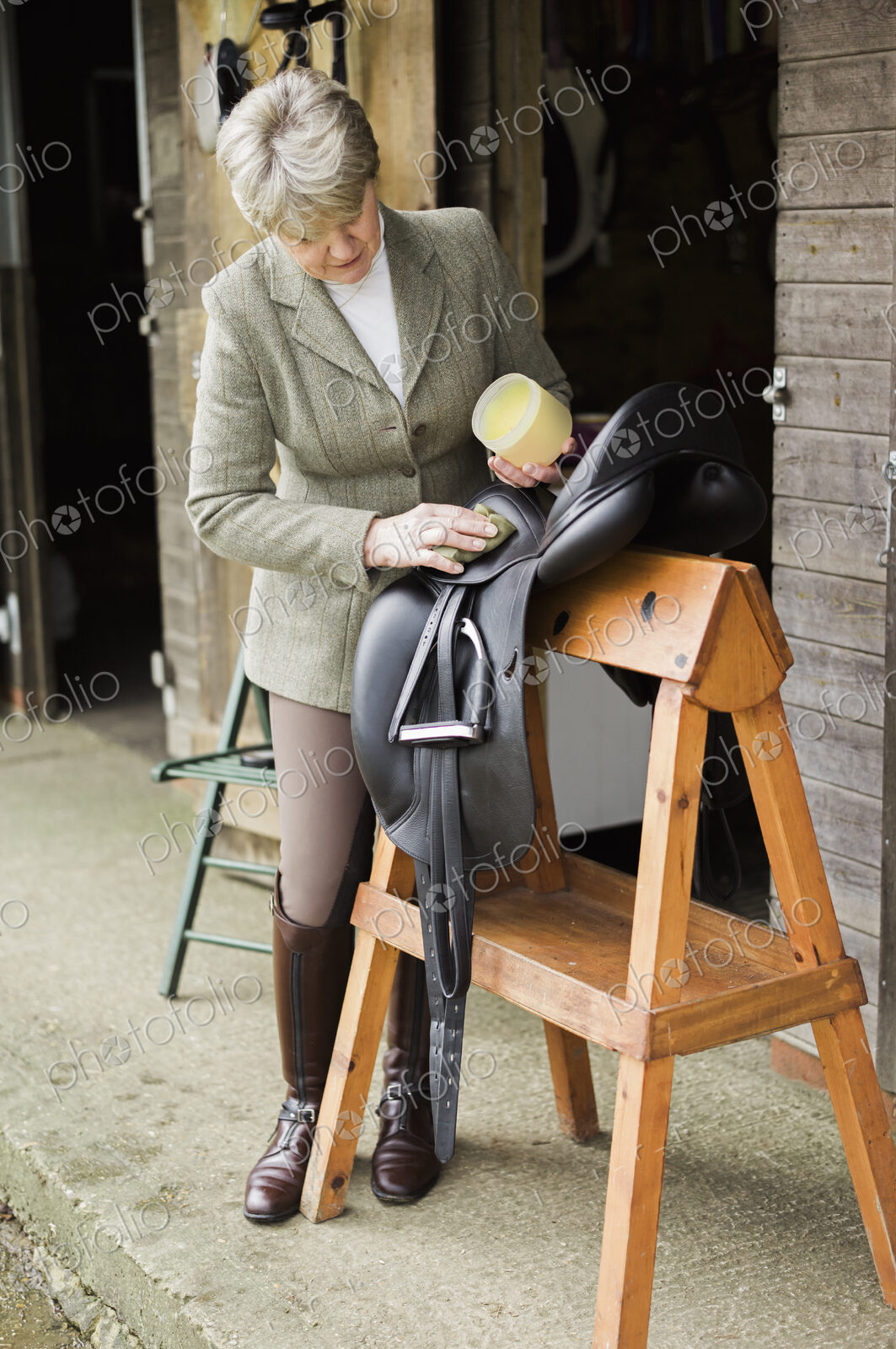 A woman cleaning and preparing tack and saddle, in the courtyard of a riding stable.