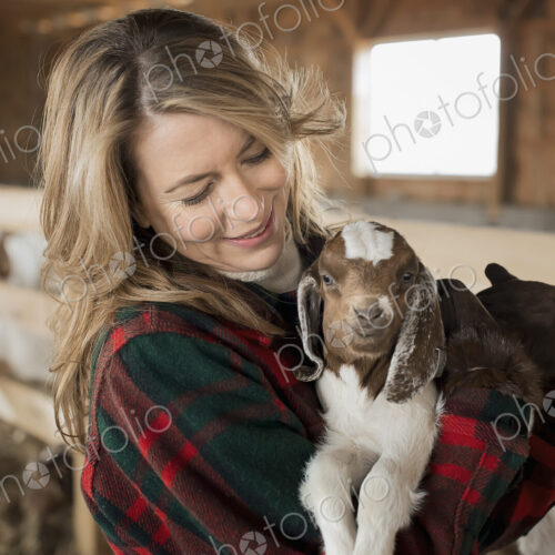 A woman cradling a young goat kid in her arms, on a farm.