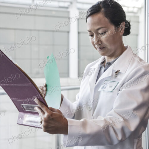 A woman doctor in a white coat, standing by a window checking a chart of patient notes. A woman doctor in a white coat, standing by a window checking a chart of patient notes.