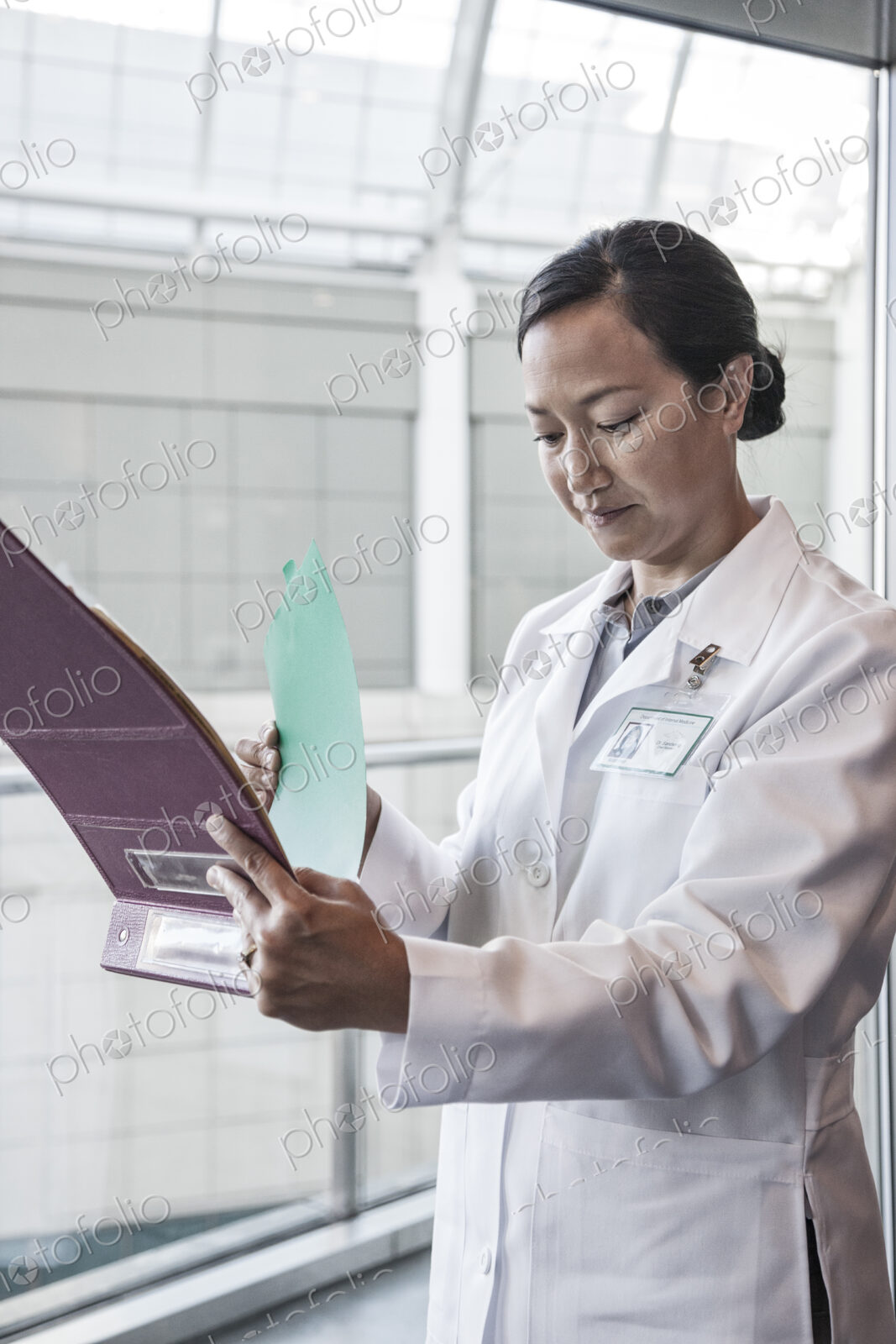 A woman doctor in a white coat, standing by a window checking a chart of patient notes.