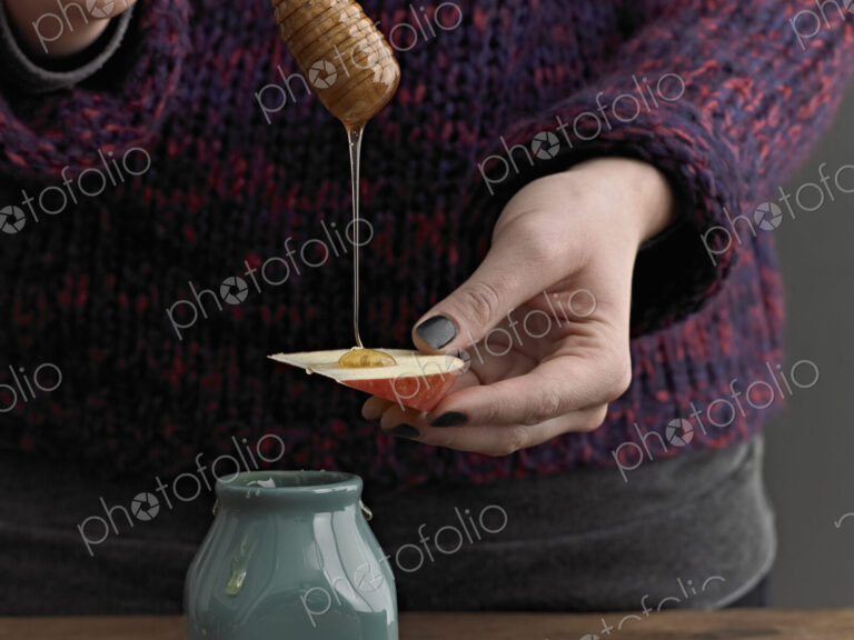 A woman drizzling honey from a round wooden drizzle stick on to an apple slice.