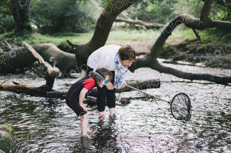 A woman giving a boy a piggyback and holding a shrimping net wading in a shallow stream.