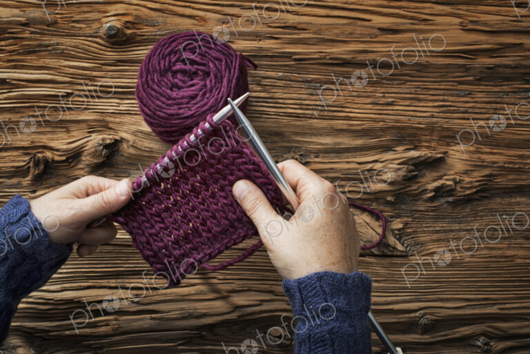 A woman holding two knitting needles, and a piece of knitting, in purple wool.