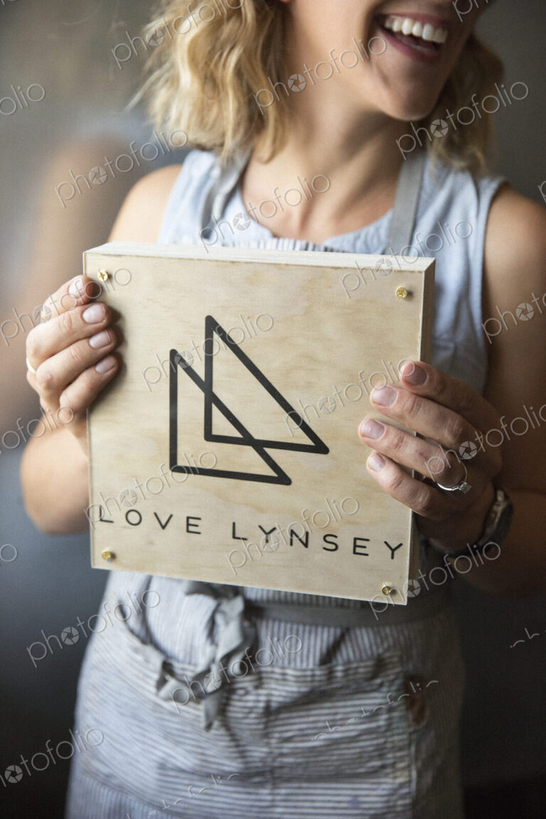 A woman in a blue apron holding her business sign, a square piece of wood with a…