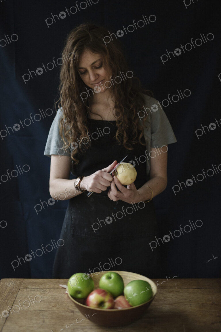 A woman in a blue apron peeling an apple with a knife.