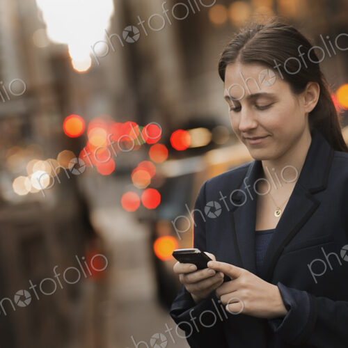 A woman in a business jacket checking her cell phone, on a city sidewalk at dusk. A woman in a business jacket checking her cell phone, on a city sidewalk at dusk.