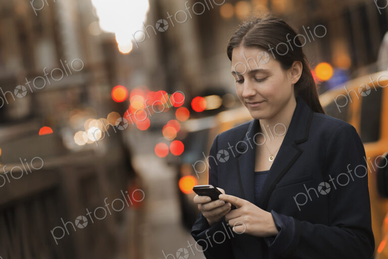 A woman in a business jacket checking her cell phone, on a city sidewalk at dusk.