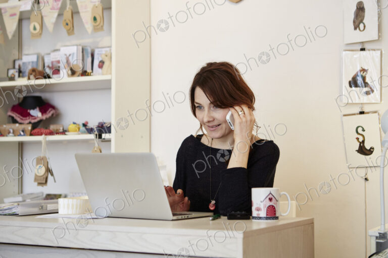 A woman in a gift shop using a laptop, making a call.