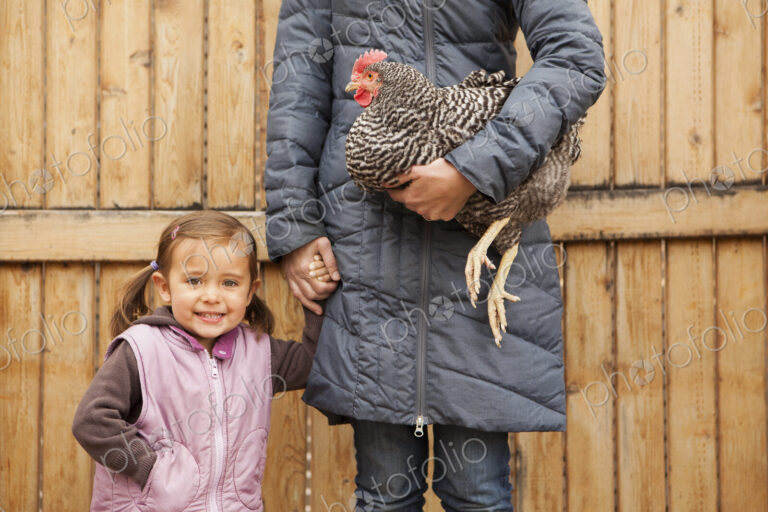 A woman in a grey coat holding a black and white chicken with a red coxcomb under one…