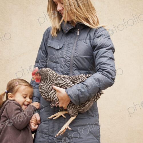 A woman in a grey coat holding a black and white chicken with a red coxcomb under one…