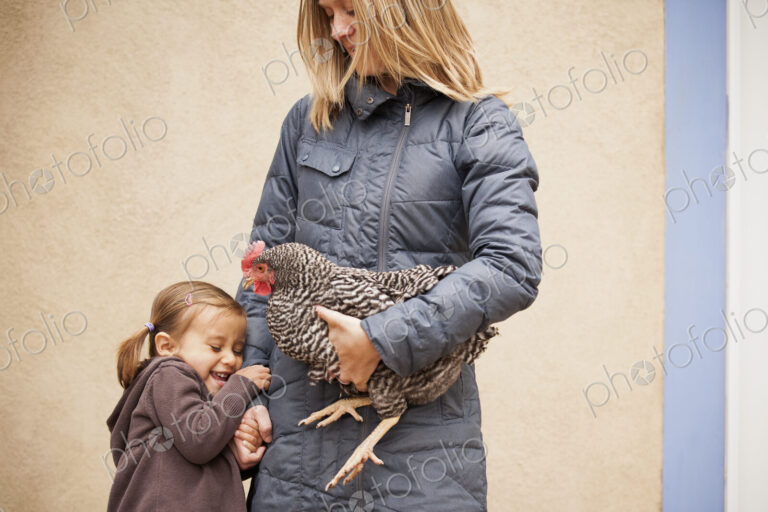A woman in a grey coat holding a black and white chicken with a red coxcomb under one…