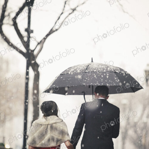 A woman in a long red evening dress and a man in a suit, walking through snow in the city.