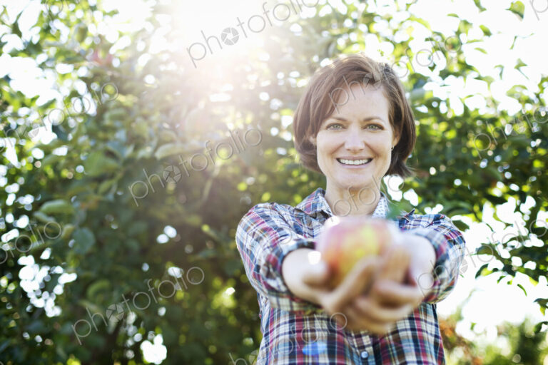 A woman in a plaid shirt holding a freshly picked apple in her two hands, in the…