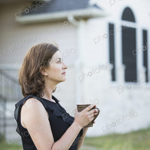 A woman in a sleeveless shirt holding a cup in her hands, standing outside a country… A woman in a sleeveless shirt holding a cup in her hands, standing outside a country…