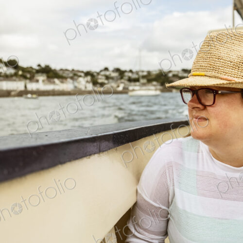 A woman in a straw hat on a boat looking out to the town on the shore on an overcast day.