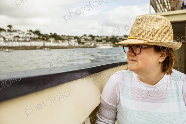 A woman in a straw hat on a boat looking out to the town on the shore on an overcast day.