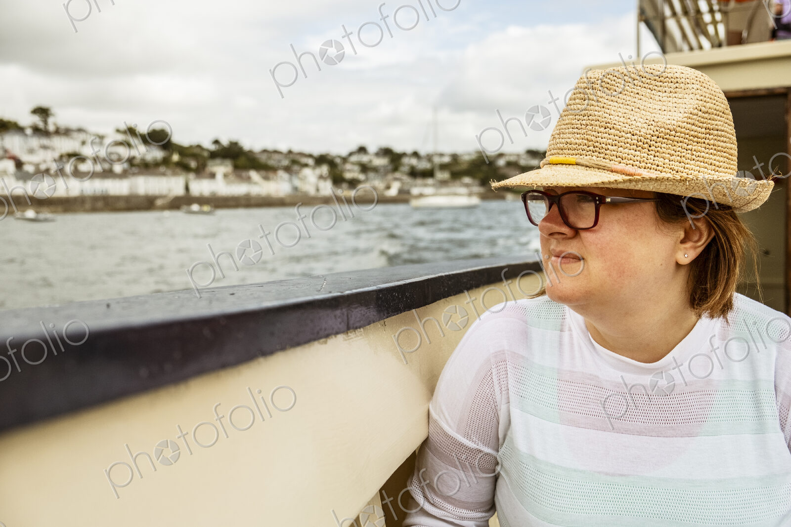 A woman in a straw hat on a boat looking out to the town on the shore on an overcast day.