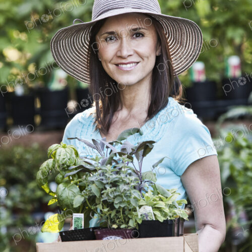 A woman in a sun hat carrying a tray of herbs and plants, shopping at a garden center…