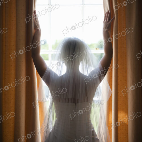 A woman in a wedding dress, wearing a light veil standing at a window with her arms up. A woman in a wedding dress, wearing a light veil standing at a window with her arms up.