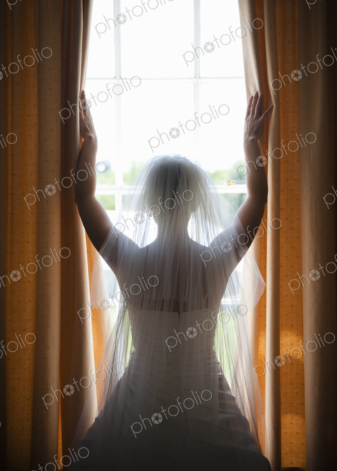 A woman in a wedding dress, wearing a light veil standing at a window with her arms up.