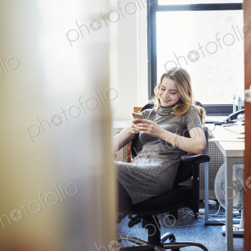 A woman in an office sitting checking her phone, seen through an open door. A woman in an office sitting checking her phone, seen through an open door.