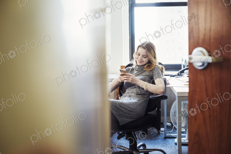 A woman in an office sitting checking her phone, seen through an open door.