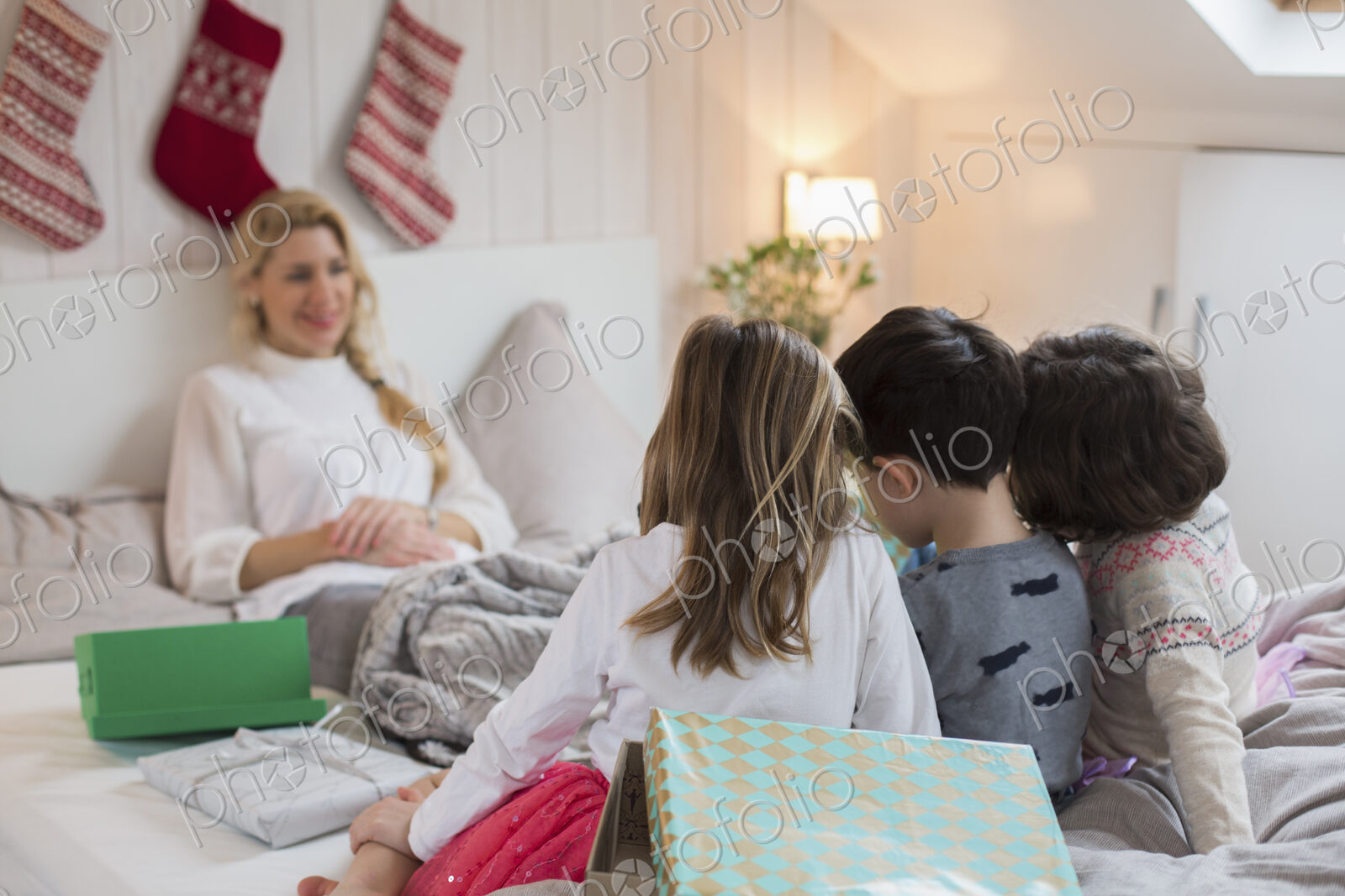 A woman in bed, with three children at the foot of the bed, carrying Christmas presents.