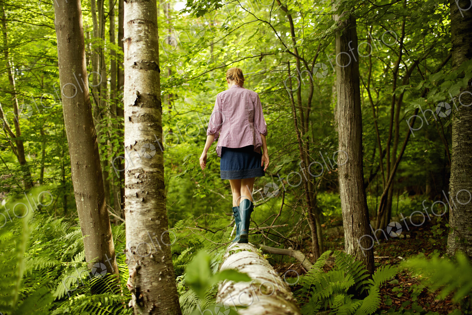 A woman in wellingtons walking along a fallen tree trunk, in woodland.